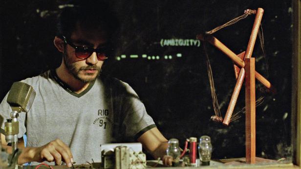 Ein Mann mit Sonnenbrille arbeitet an einer Apparatur mit Röhren und einem Holzmodell vor einer Tafel mit Morsecode.