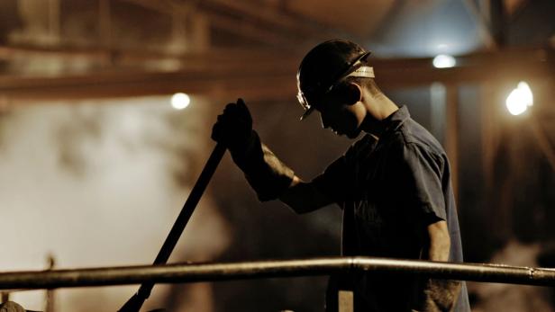 Ein Arbeiter mit Schutzhelm und Brille hantiert mit einem Werkzeug in einer Fabrikhalle.