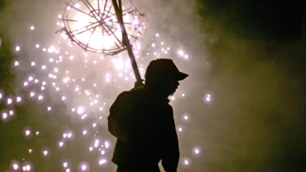 Eine Person in Silhouette trägt ein Feuerwerksrad auf einer Stange.