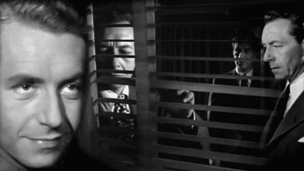 Drei Männer blicken durch Jalousien in einem Schwarzweißfilm.