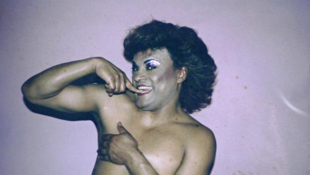 Eine barbusige Person mit Make-up posiert vor einem rosa Hintergrund.