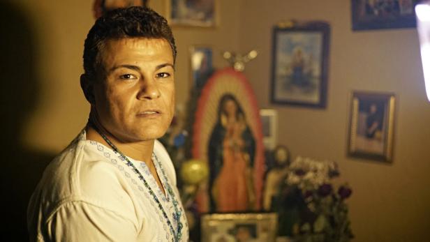 Ein Mann mit Halskette steht vor einem Wandaltar mit religiösen Bildern.