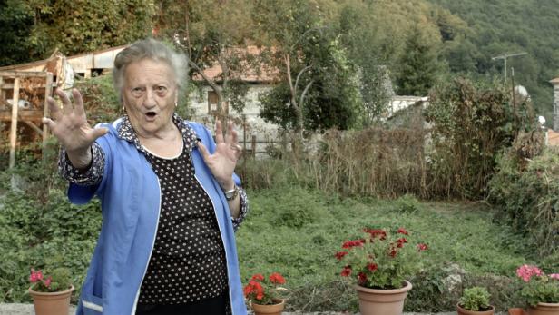 Eine ältere Frau gestikuliert vor einem Garten mit Blumen.
