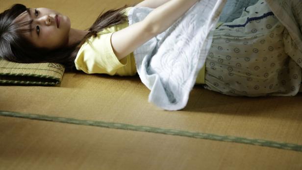 Eine junge Frau liegt auf einer Tatami-Matte und deckt sich mit einer hellblauen Decke zu.