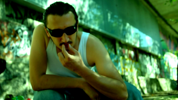 Ein Mann mit Sonnenbrille und Schnurrbart isst eine rote Beere vor einer Graffitiwand.