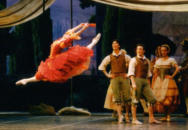 Eine Ballerina in einem roten Tutu springt auf einer Bühne, während andere Tänzer zusehen.