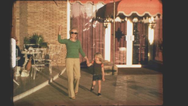 Eine Frau winkt, während sie mit einem Kind vor einem Gebäude entlanggeht.