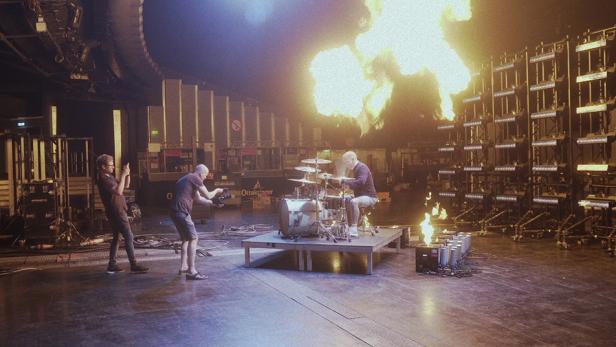 Ein Schlagzeuger spielt vor einer Feuerwand auf einer Bühne, während zwei Personen filmen.