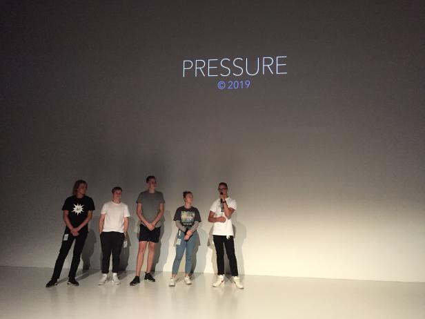 Fünf Personen stehen vor einer Leinwand mit der Aufschrift „Pressure © 2019“.