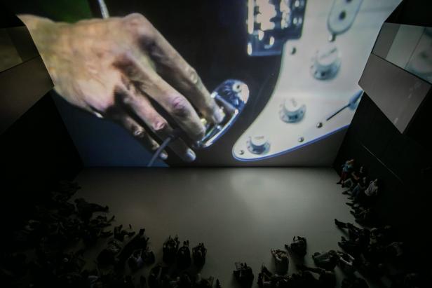 Ein Publikum schaut auf eine große Projektion einer Hand auf einer Gitarre.