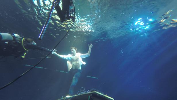 Ein Mann steht unter Wasser auf einer Plattform, während Taucher ihn sichern.