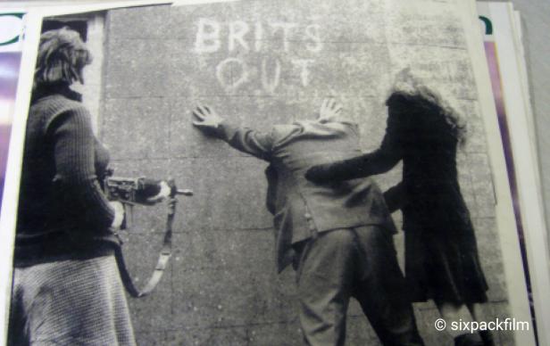 Eine Frau mit Gewehr bewacht einen Mann, der an einer Wand mit der Aufschrift „Brits Out“ steht.
