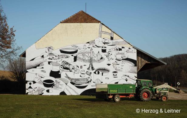 Ein Bauernhaus mit einem großen Wandgemälde, das ein Festmahl darstellt, und einem Traktor davor.