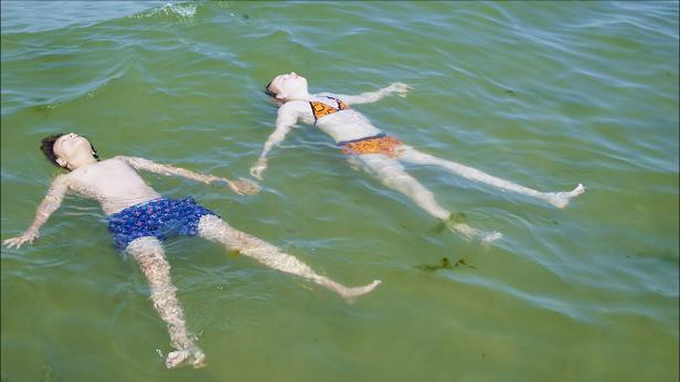 Zwei Kinder treiben entspannt im flachen, grünen Wasser.