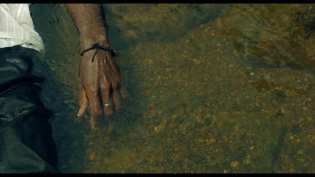 Eine Hand mit Armband und Ring berührt das klare Wasser eines Baches.