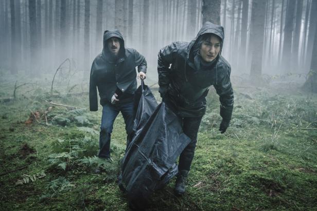 Zwei Personen tragen eine schwarze Plane durch einen nebligen Wald.