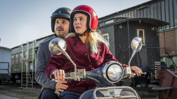 Ein Paar mit Helmen fährt auf einem blauen Motorroller vor alten Eisenbahnwaggons.