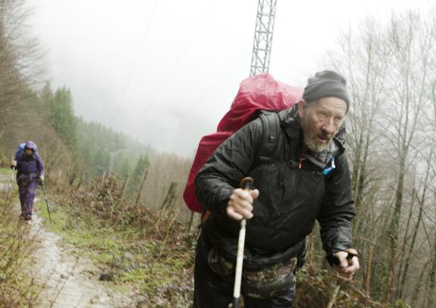 Ein Mann wandert mit einem roten Rucksack und Wanderstöcken auf einem schlammigen Pfad.