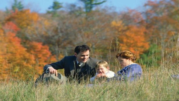 Eine Familie sitzt in einem Feld mit herbstlichen Bäumen im Hintergrund.