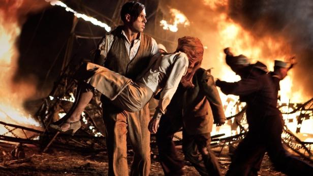 Ein Mann trägt eine bewusstlose Frau vor einem brennenden Hintergrund davon.