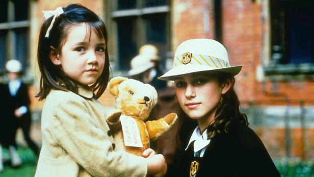Zwei junge Mädchen, eines mit einem Teddybär, stehen vor einem Backsteingebäude.