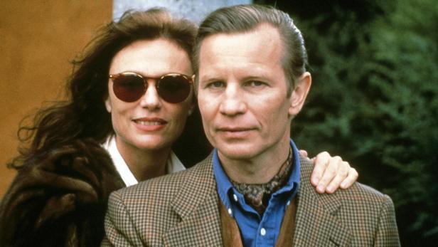 Ein Mann im Tweed-Sakko mit Krawatte und eine Frau mit Sonnenbrille posieren für ein Foto.