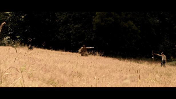 Drei Männer stehen in einem hohen Grasfeld vor einem dunklen Wald.