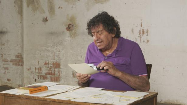 Ein Mann sitzt an einem Tisch und liest ein Dokument mit einer Lupe. Auf dem Tisch liegen weitere Dokumente und ein Buch.