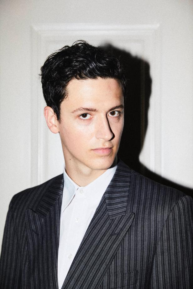 Ein junger Mann mit dunklen Haaren trägt ein gestreiftes Jackett und ein weißes Hemd.