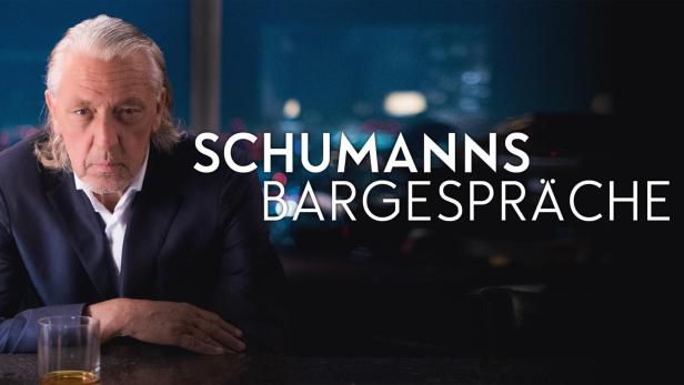 Ein älterer Mann im Anzug sitzt an einer Bar mit dem Schriftzug „Schumanns Bargespräche“.