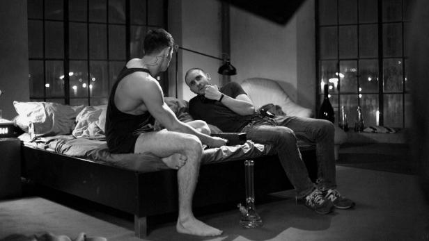 Zwei Männer sitzen in einem Zimmer auf einem Bett und unterhalten sich.