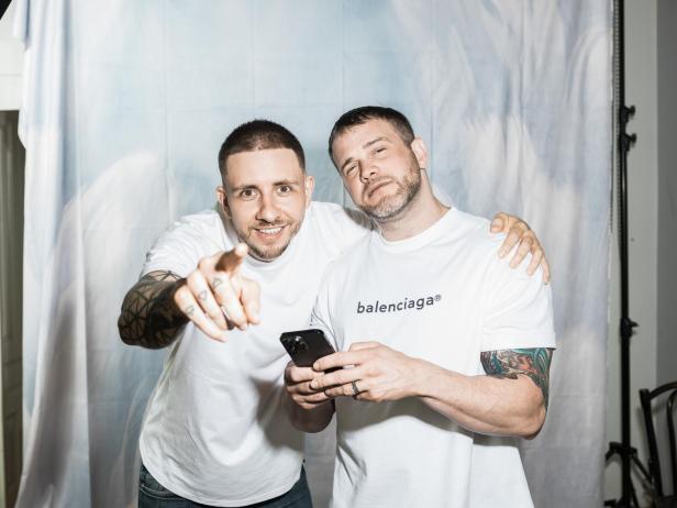 Zwei Männer mit Tattoos posieren, einer zeigt mit dem Finger in die Kamera, der andere hält ein Smartphone.