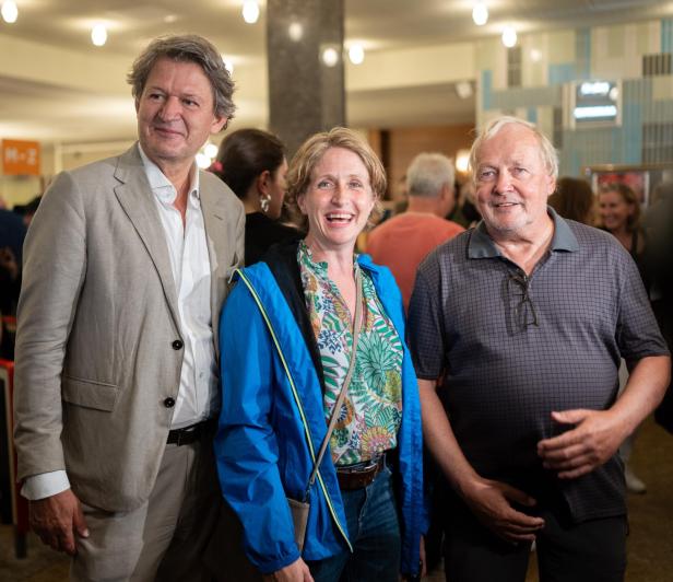 Drei Personen stehen lächelnd in einem belebten Raum.