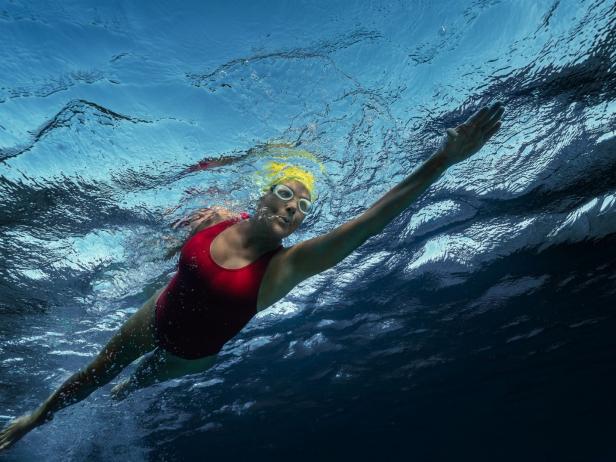 Eine Frau mit gelber Badekappe und rotem Badeanzug schwimmt unter Wasser.
