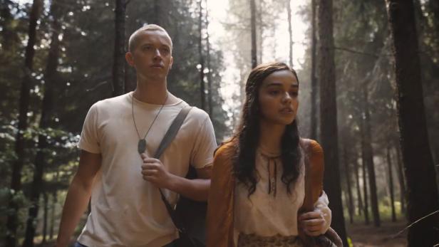 Tom Blyth mit Army-Kette und weißem T-Shirt mit Rachel Zegler im Wald