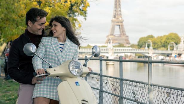 Ein Mann und eine Frau lächeln sich verliebt auf einer Vespa an der Seine an. Hinter ihnen sieht man den Eiffelturm.