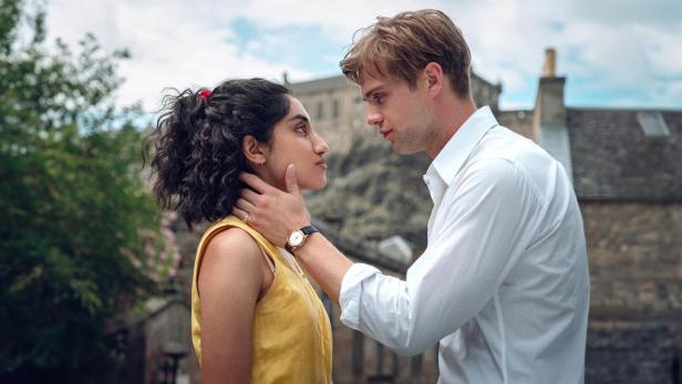 Ein weißer, blonder, junger Mann in einem weißen Hemd legt seine linke Hand liebevoll an den Hals einer jungen Frau mit indischer Herkunft, die ein gelbes T-Shirt trägt.