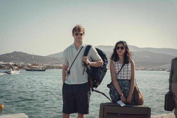 Ein blonder, junger Mann und eine junge Frau mit indischen Wurzeln stehen mit Sonnenbrillen an einem Hafen vor einer griechischen Insel.