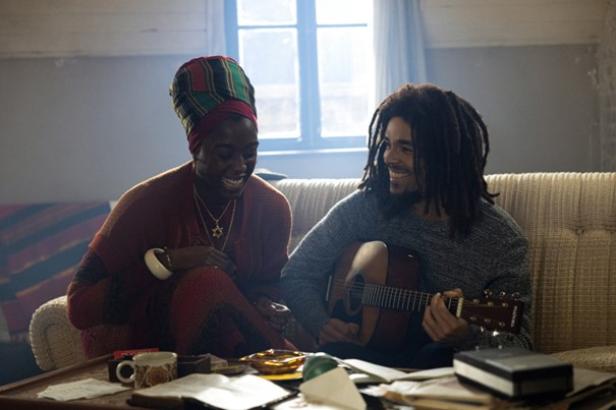 Eine Schwarze Frau mit einem bunten Turban sitzt neben einem Schwarzen Mann mit Rastalocken, der Gitarre spielt. Beide lächeln.