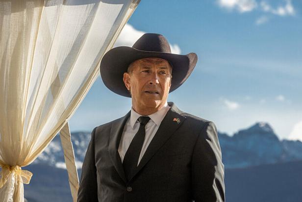 Ein Mann steht im Anzug und einem Cowboyhut vor einer Bergkulisse.