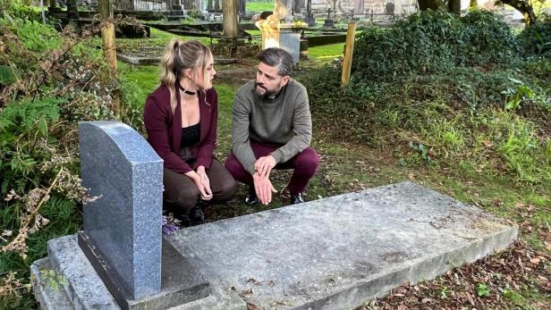 Ein Mann und eine Frau kauern an einem Grabstein auf einem Friedhof und unterhalten sich.