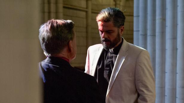 Ein bärtiger Mann im Priesterhemd und hellem Sakko unterhält sich mit einer anderen Person.