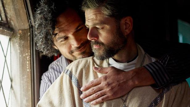 Ein Mann umarmt einen anderen Mann mit Bart vor einem Fenster.