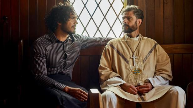 Ein Mann im gestreiften Hemd sitzt neben einem Priester in einer Kirche.