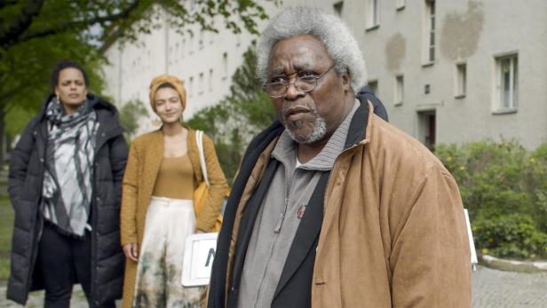 Ein älterer Mann mit Brille steht vor einem Gebäude, begleitet von zwei Frauen.