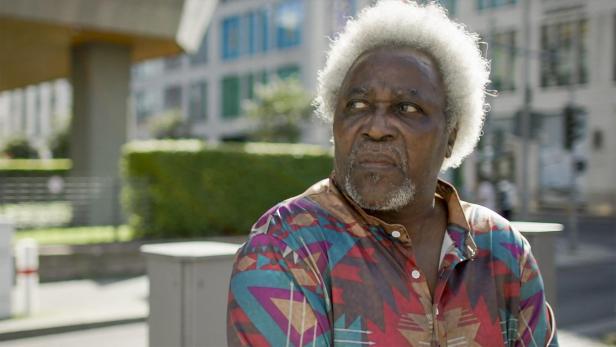 Ein älterer, dunkelhäutiger Mann mit weißem Haar und buntem Hemd blickt nach links.