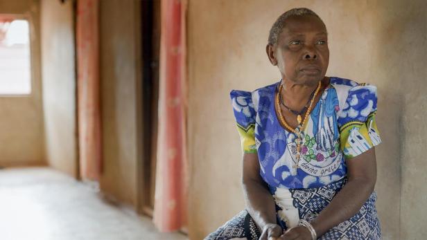 Eine ältere Frau mit Halsketten sitzt in einem Raum und blickt nachdenklich zur Seite.