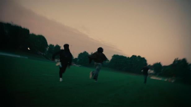 Drei Personen rennen bei Dämmerung über eine grüne Wiese.