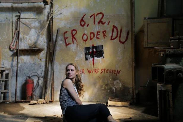 Eine Frau sitzt in einem heruntergekommenen Raum mit der Aufschrift „6.12. Er oder Du. Er wird sterben“ an der Wand.