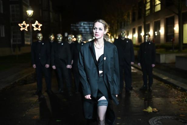 Eine Frau in schwarzem Blazer geht vor einer Gruppe maskierter Männer auf der Straße.